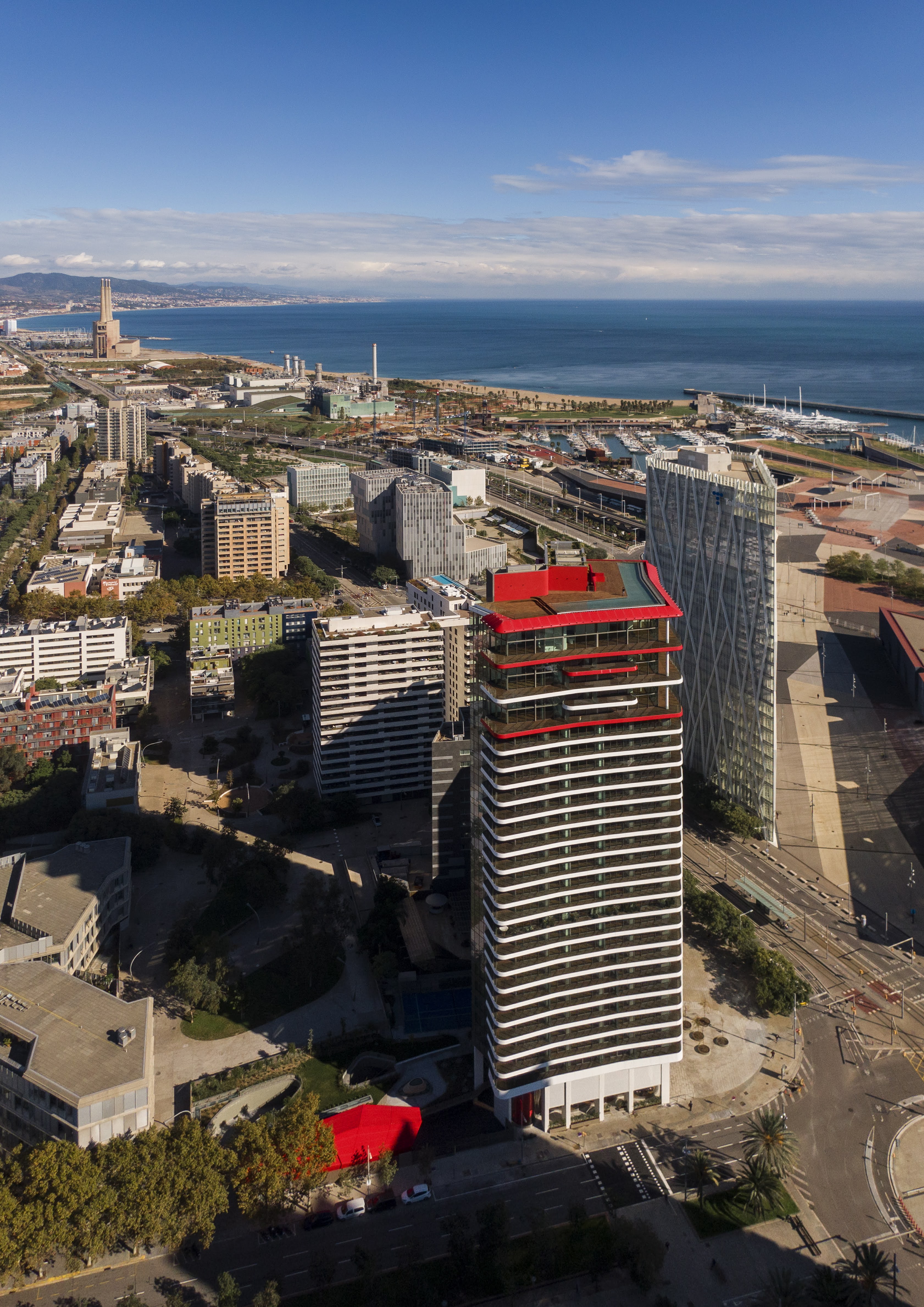 High-angle aerial view of the Antares Barcelona residential tower with its signature red roof and curved terraces, located in the Diagonal Mar tech district near the Mediterranean coast.