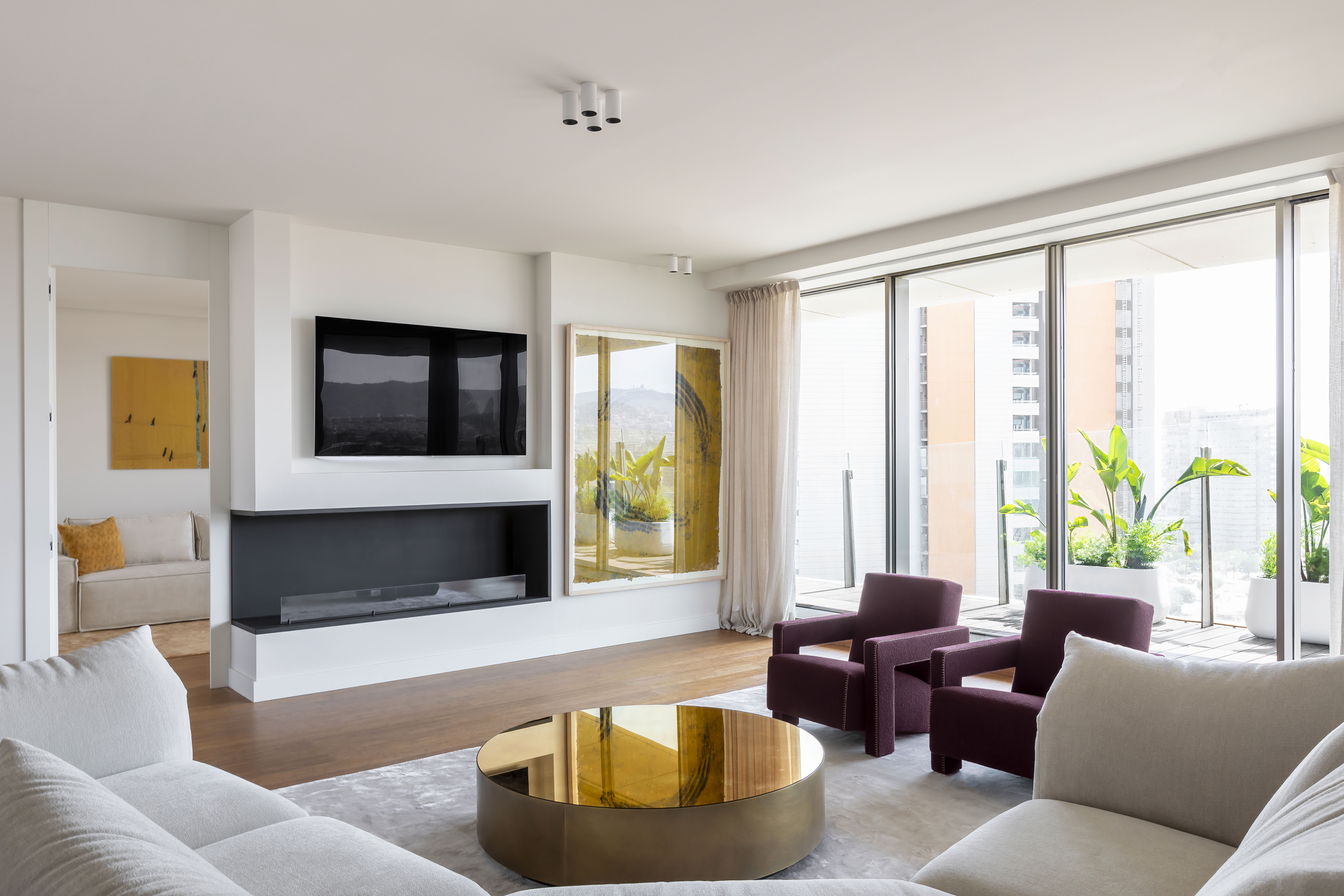 Modern minimalist living room in an Antares Barcelona apartment featuring a gold cylindrical coffee table, deep burgundy velvet armchairs, and a sleek recessed fireplace against a white gallery-style wall.