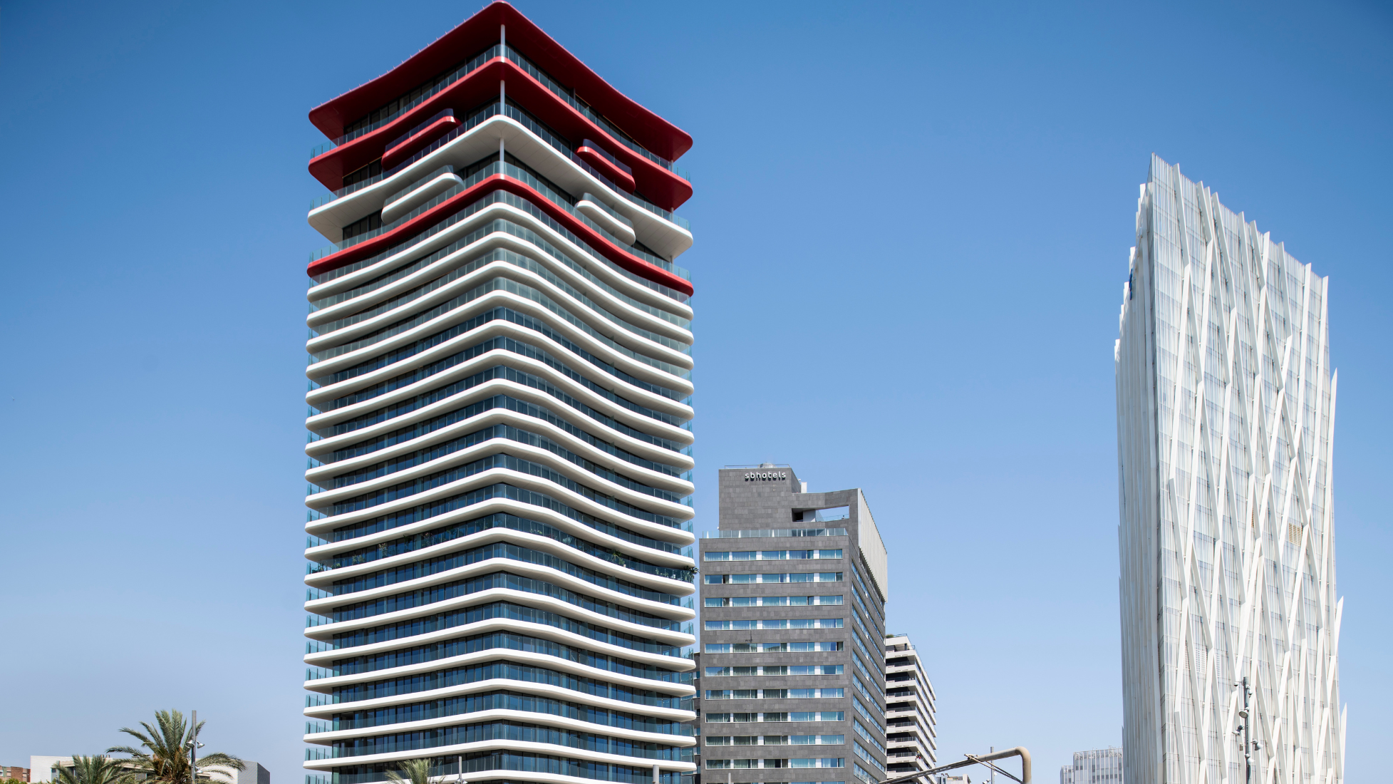The exterior of the Antares tower in Barcelona, characterized by its undulating white balconies and bold red top floors. The sculptural building stands as a modern landmark against a clear blue sky in the Diagonal Mar district.
