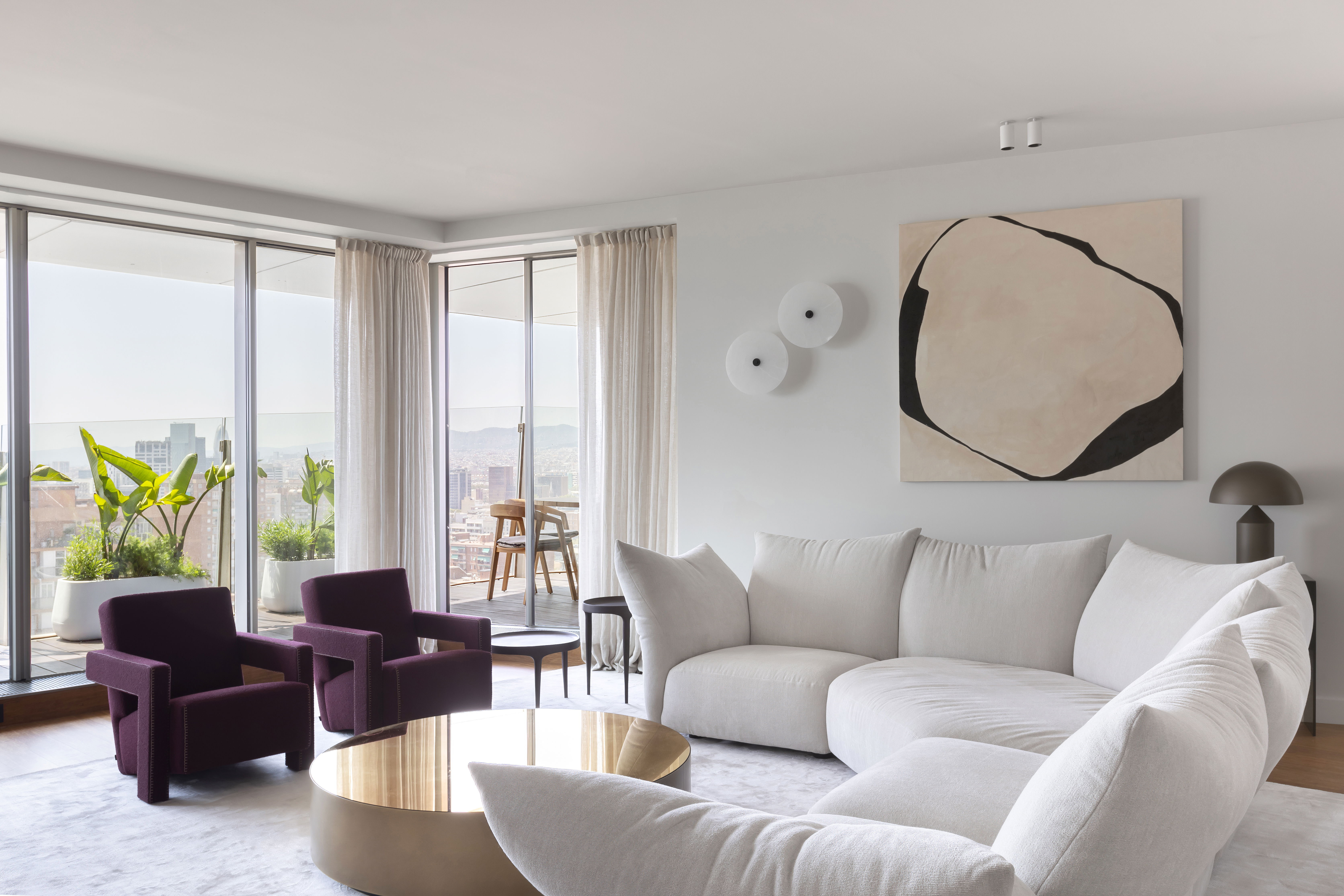 A panoramic view of an Antares Barcelona living room featuring a curved white sectional sofa, a gold reflective coffee table, and plum-colored accent chairs against floor-to-ceiling windows overlooking the city.