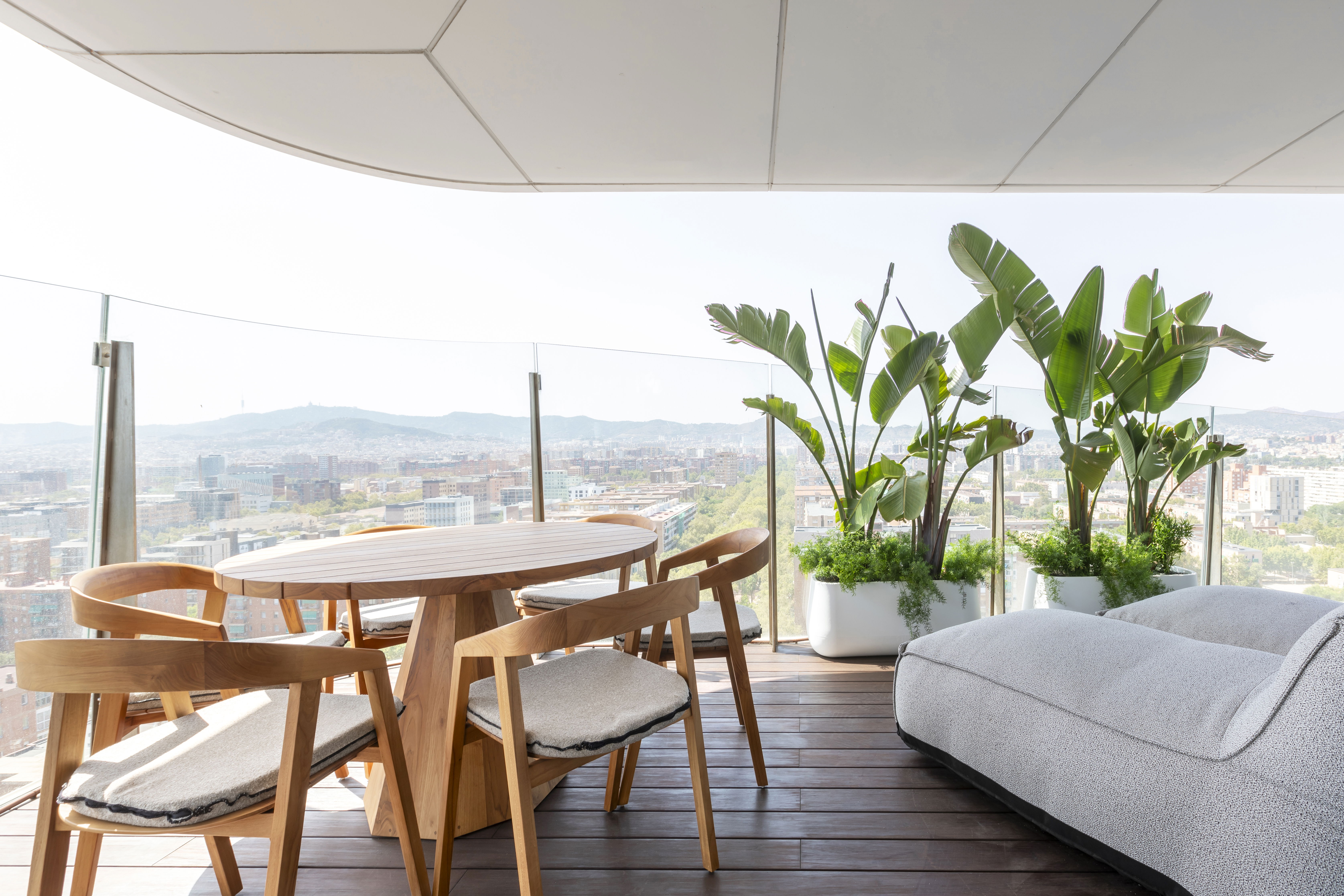 A high-floor private terrace at Antares Barcelona featuring a round wooden dining table, modern chairs, lush potted tropical plants, and a panoramic view of the city.
