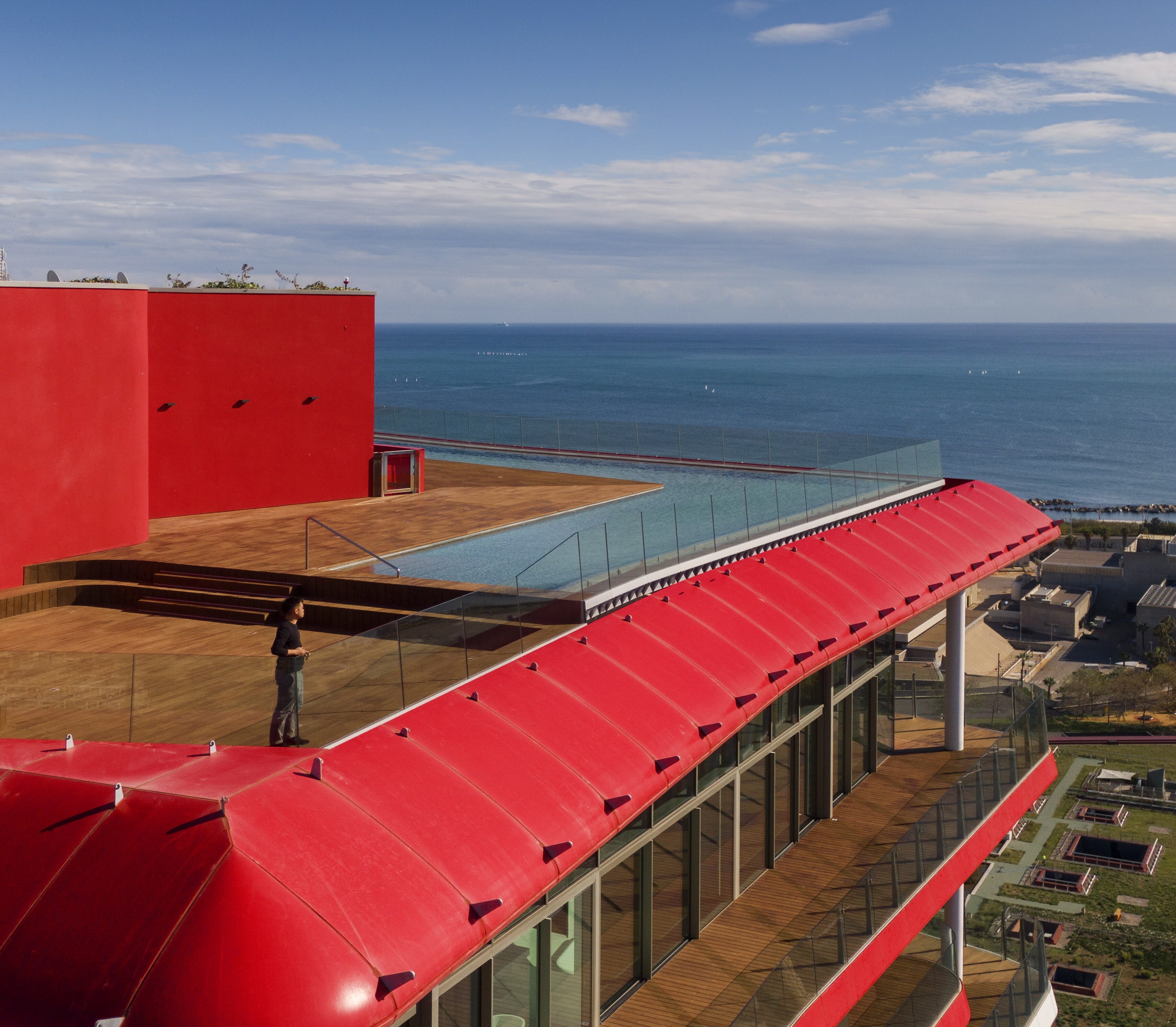 High-angle exterior shot of Antares Barcelona’s iconic red rooftop terrace and infinity pool overlooking the Mediterranean Sea, highlighting the bold architecture of a premium real estate investment.