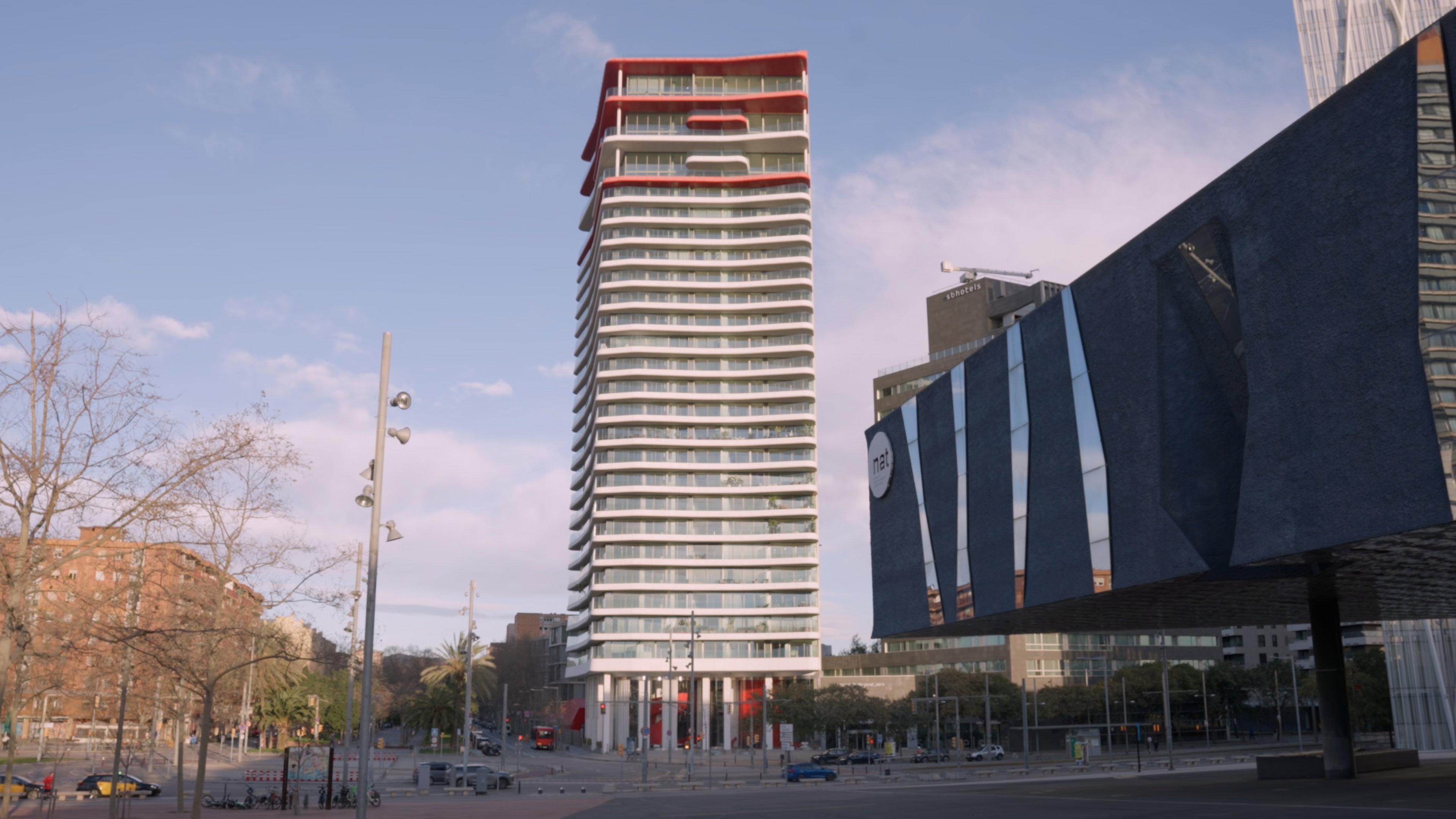 A wide-angle exterior view of Antares Barcelona, a high-rise luxury residential tower featuring distinctive red-and-white undulating balconies. The building is positioned as an architectural landmark and secure urban retreat in the Diagonal Mar district, shown alongside the modern Museu de Ciències Naturals under a clear sky.