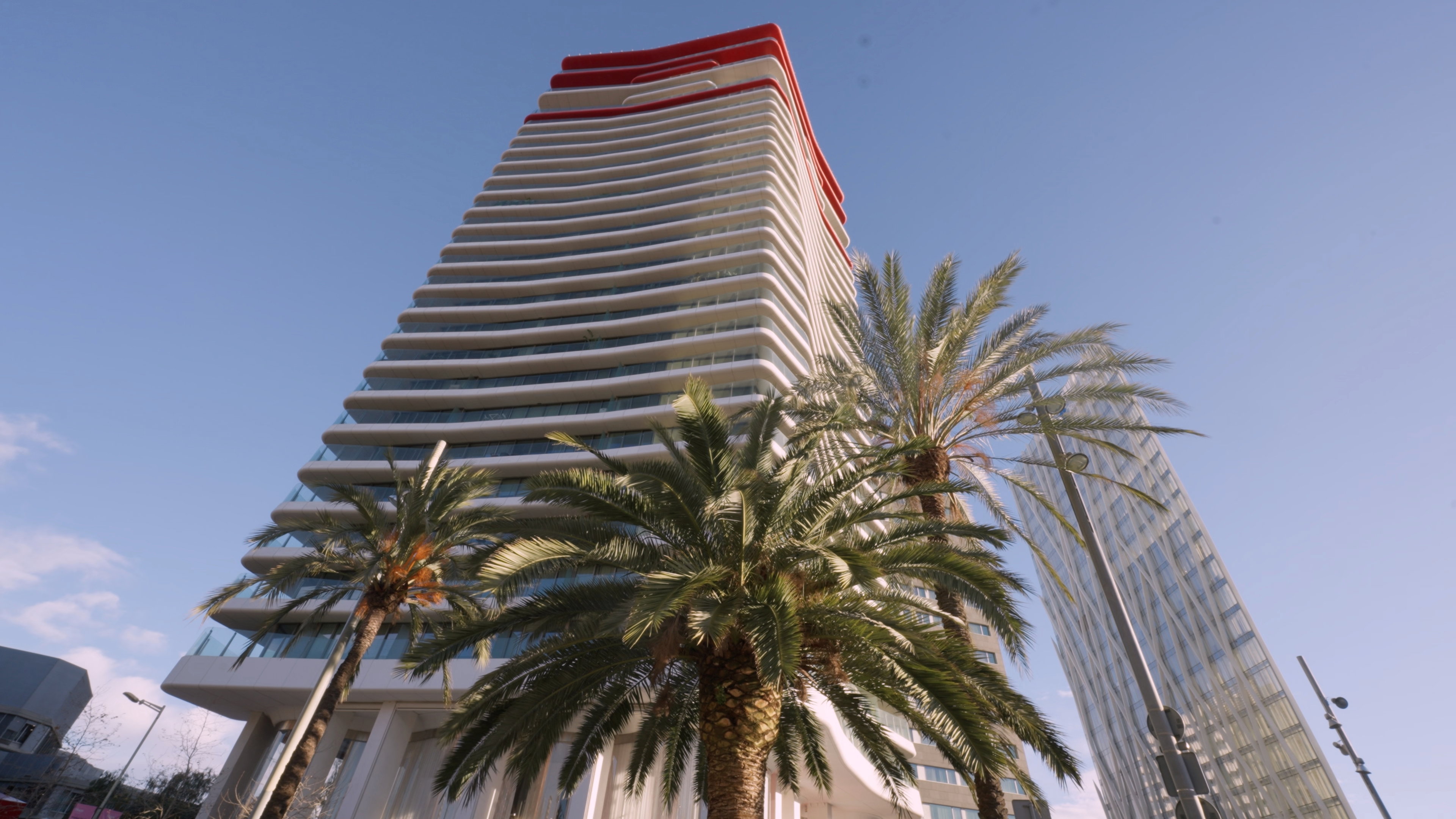 Low-angle perspective of the Antares Barcelona residential tower, highlighting its undulating white balconies and distinct red roof against a clear blue sky. Two prominent palm trees frame the architecture, symbolizing the city's Mediterranean luxury lifestyle, with the glass facade of the Torre Diagonal ZeroZero visible in the background as a marker of modern investment.