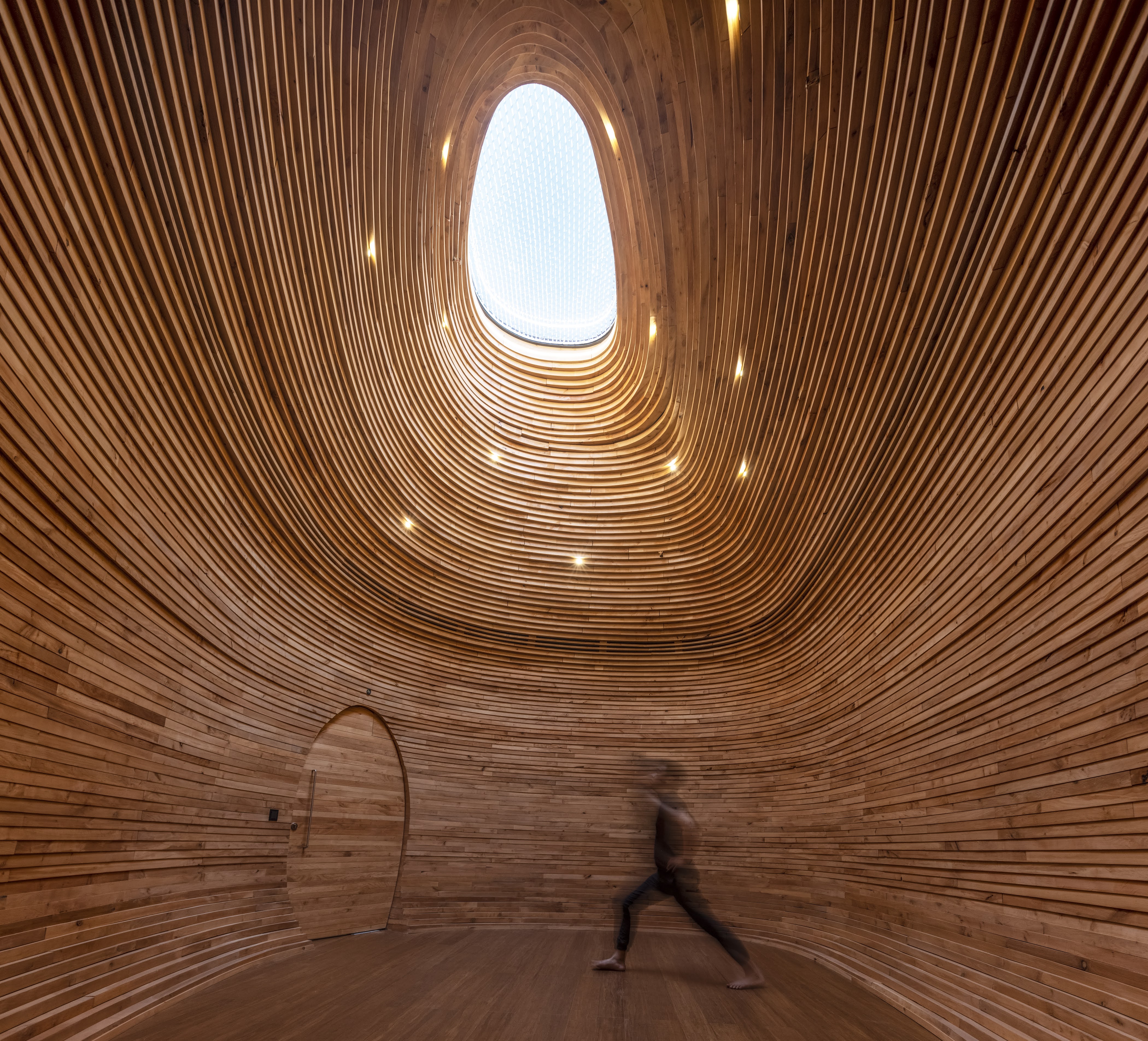 Interior view of the luxury yoga space inside Antares Barcelona, featuring an organic, cocoon-like room lined with undulating horizontal wooden slats, a curved wooden door, and a striking, asymmetrical skylight funneling natural light from above.
