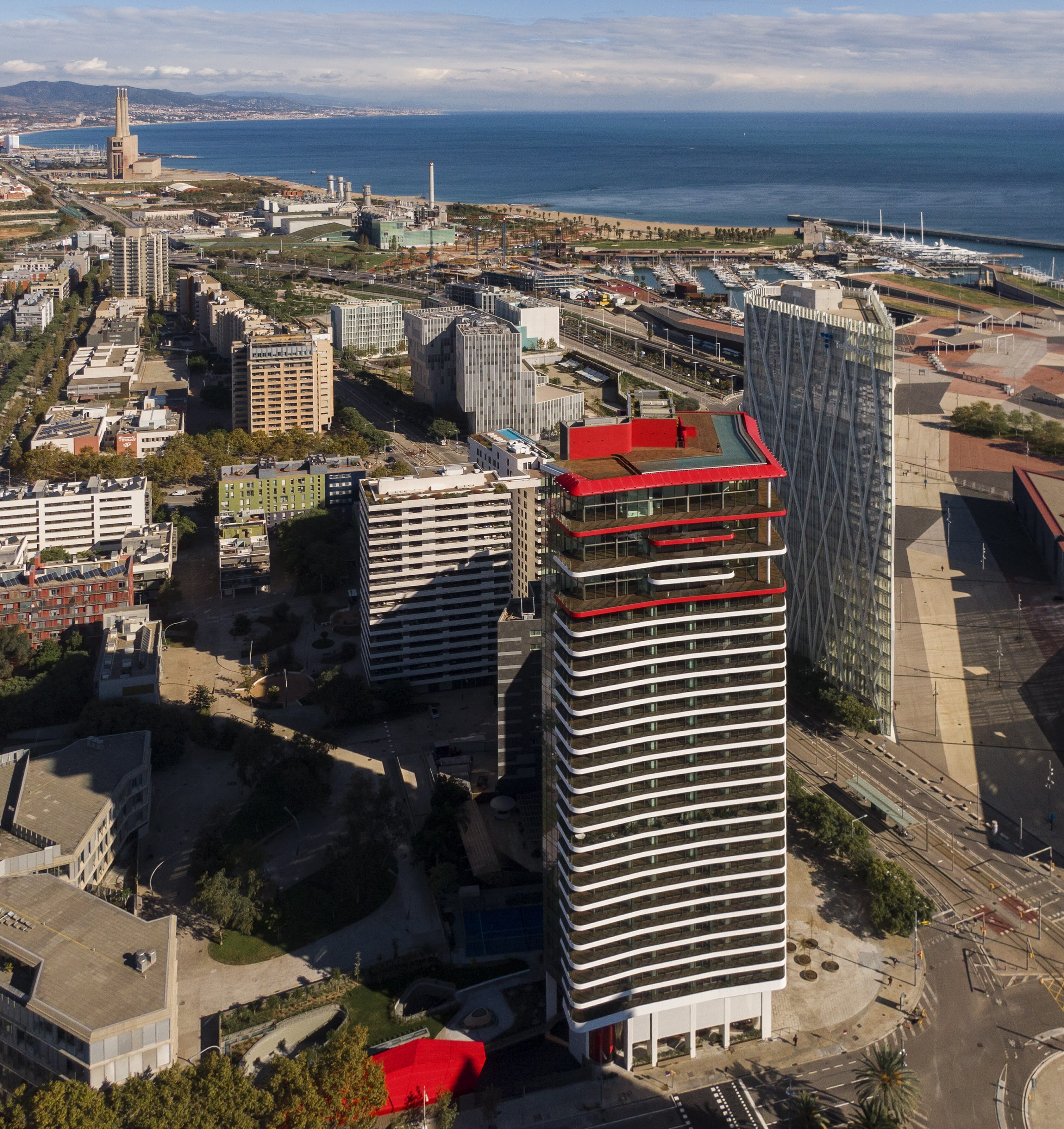High-angle aerial view of Barcelona's modern Diagonal Mar and Forum district along the coast, showcasing contemporary high-rise towers, the distinctive red-accented Antares building, and the deep blue Mediterranean Sea stretching out to the horizon.