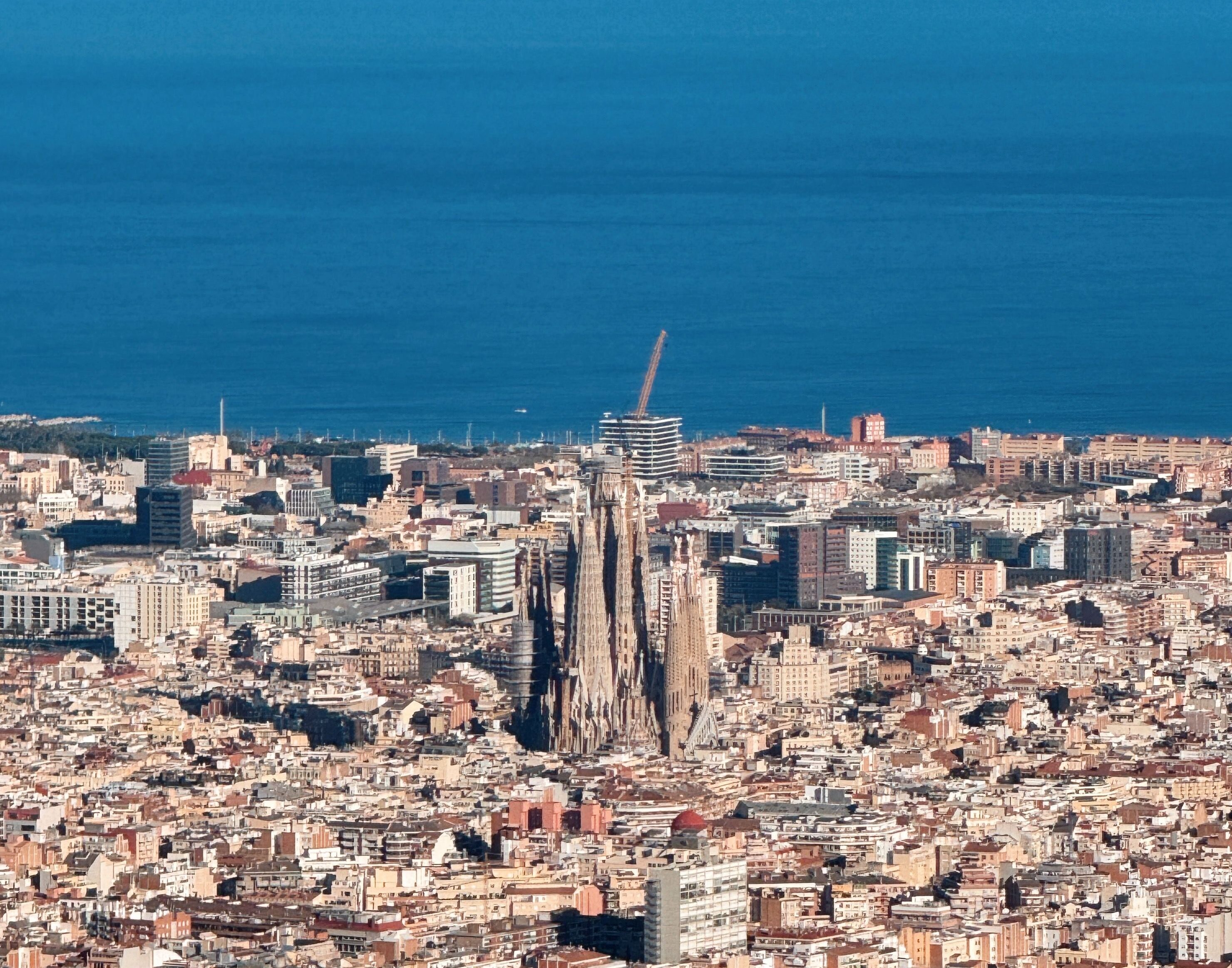 Panoramic high-angle view of the dense Barcelona cityscape stretching out to the deep blue Mediterranean Sea, with the towering spires of Antoni Gaudí's Sagrada Família standing out prominently in the center.