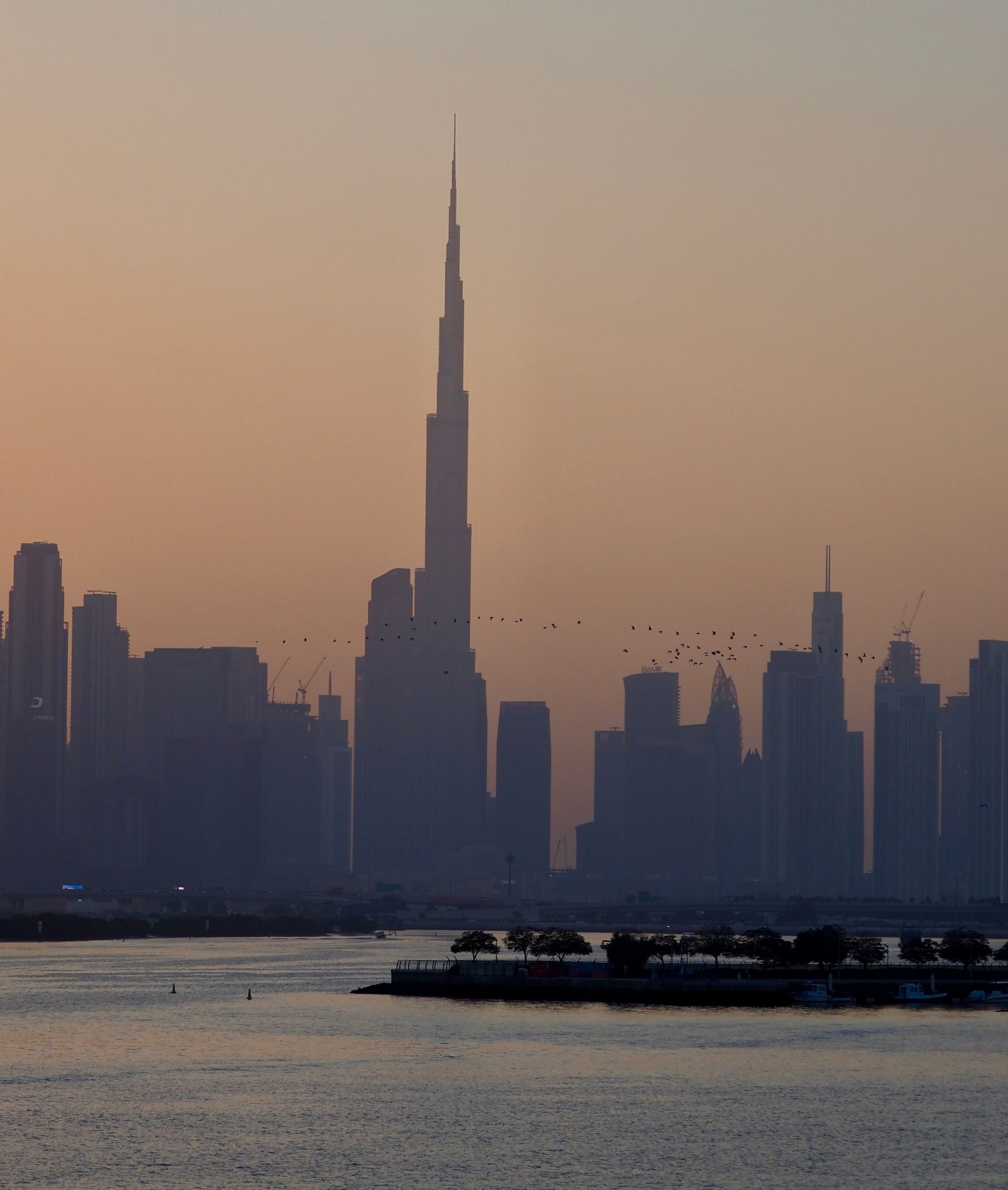 distant view across the water to the silhouette of the Burj Khalifa and the Dubai skyline at sunset. A flock of birds flies in the sky. This represents the wealth and infrastructure of Dubai before a narrative shift to Barcelona.