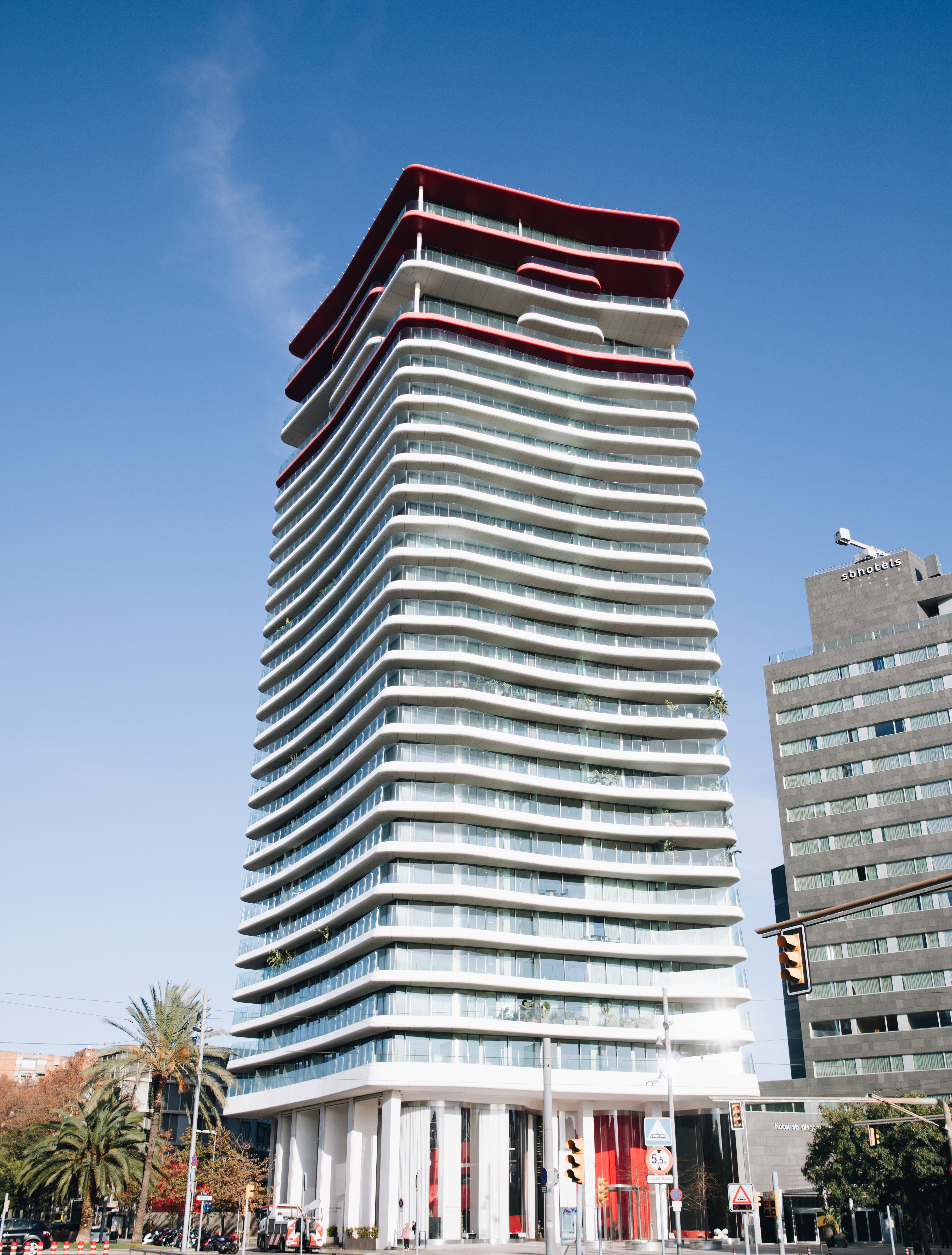 Exterior low-angle view of a tall, modern residential skyscraper featuring distinctive white curved balconies and red accents on top against a clear blue sky.
