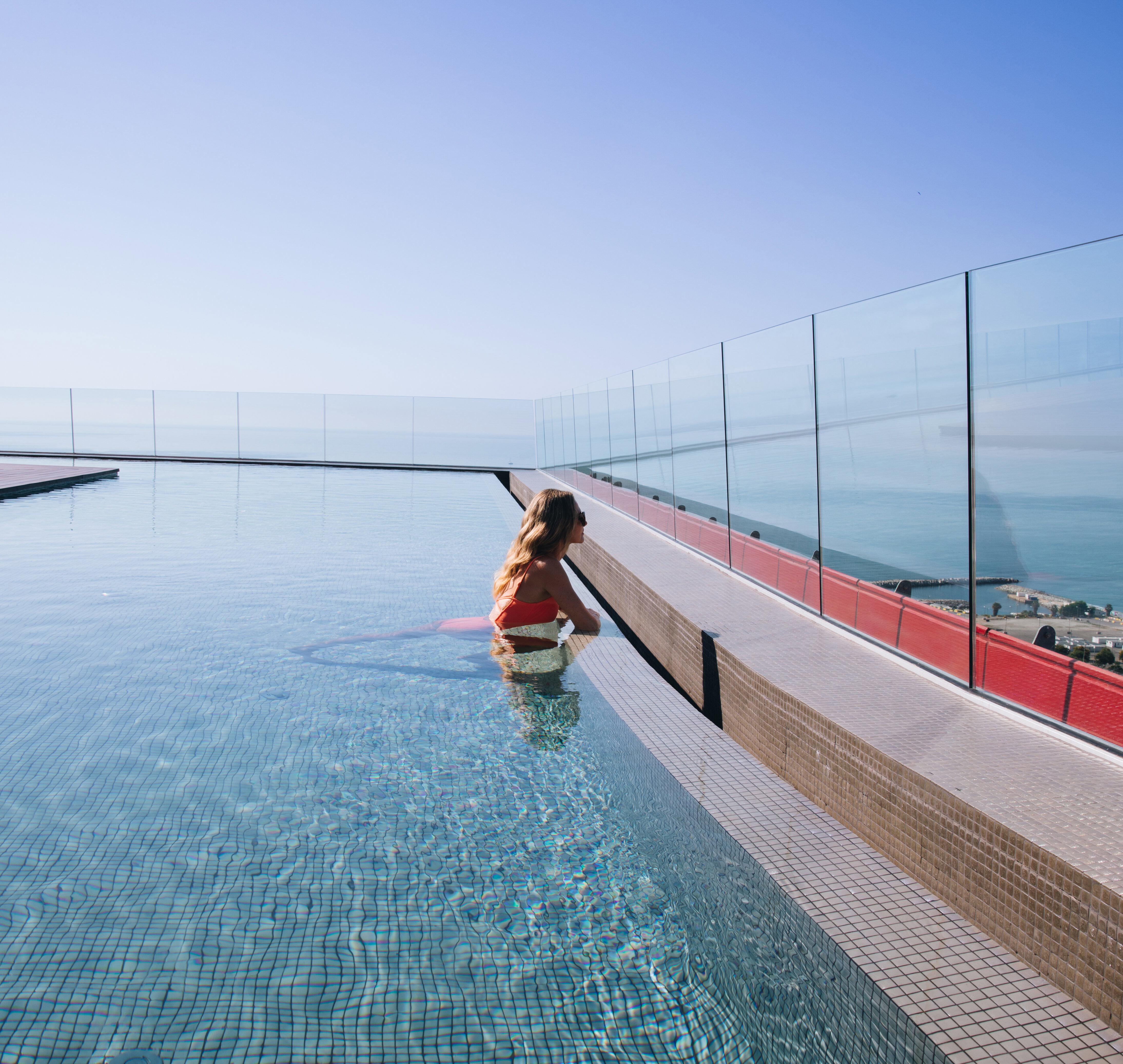 A woman in an orange swimsuit rests at the edge of a high-rise rooftop swimming pool enclosed by glass walls, gazing out toward the calm ocean horizon under a clear blue sky.